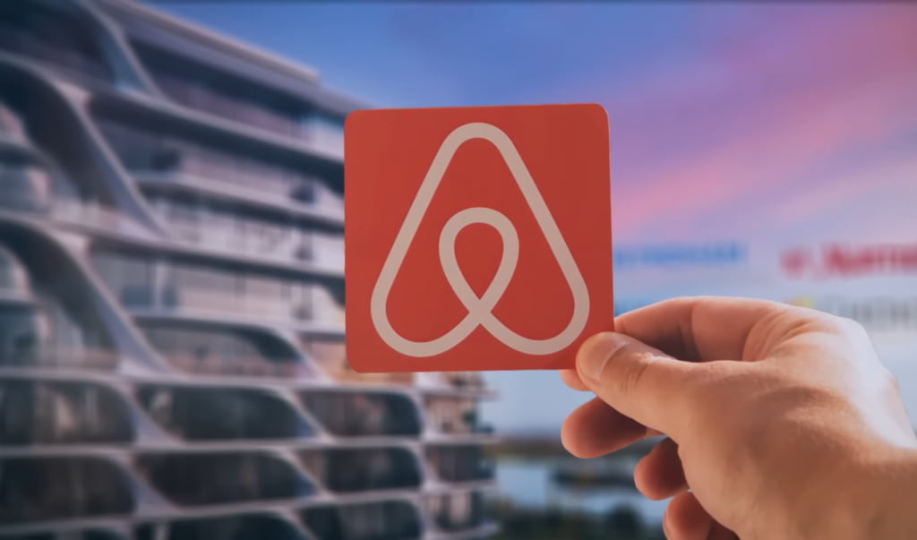 A hand holds up a red card with a white Airbnb logo, with a cityscape in the background