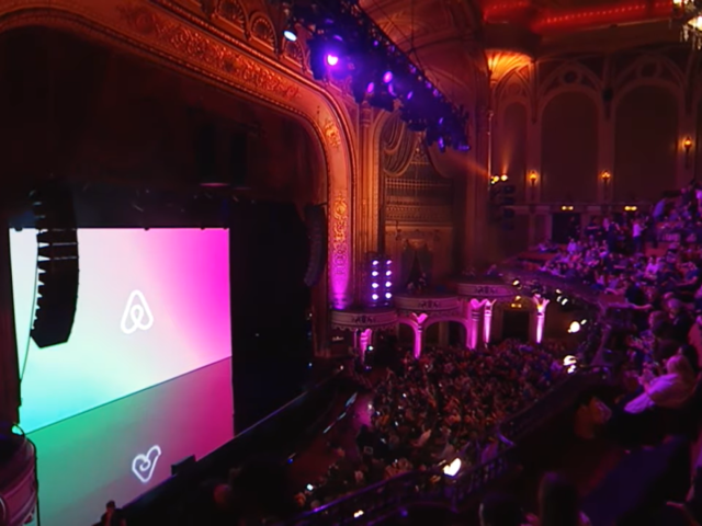 A grand theater lit in pink, with Airbnb's logo on screen and an eager audience awaiting the event