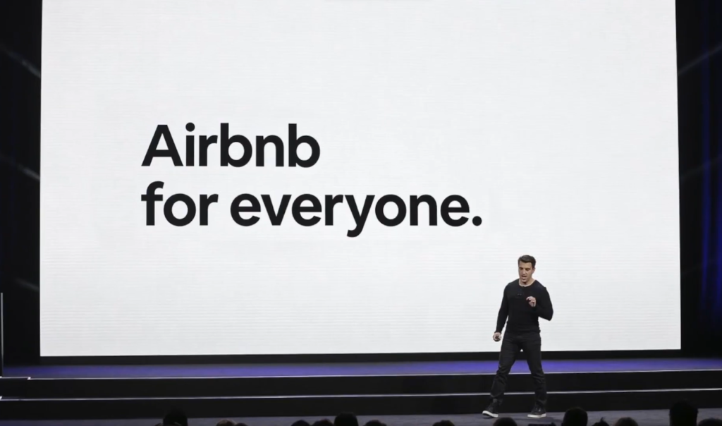 A man presents on stage with "Airbnb for everyone." displayed on a large screen behind him