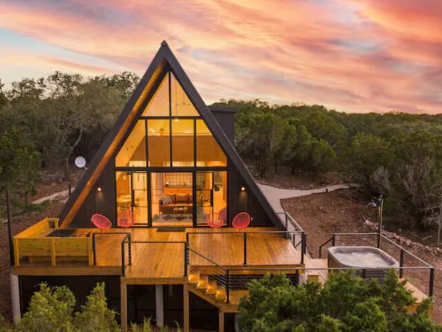 A-frame house with large windows, a wooden deck, and a hot tub, set against a sunset sky.