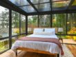 A bedroom with glass walls and ceiling in a forest setting, with a bed and tree artwork.